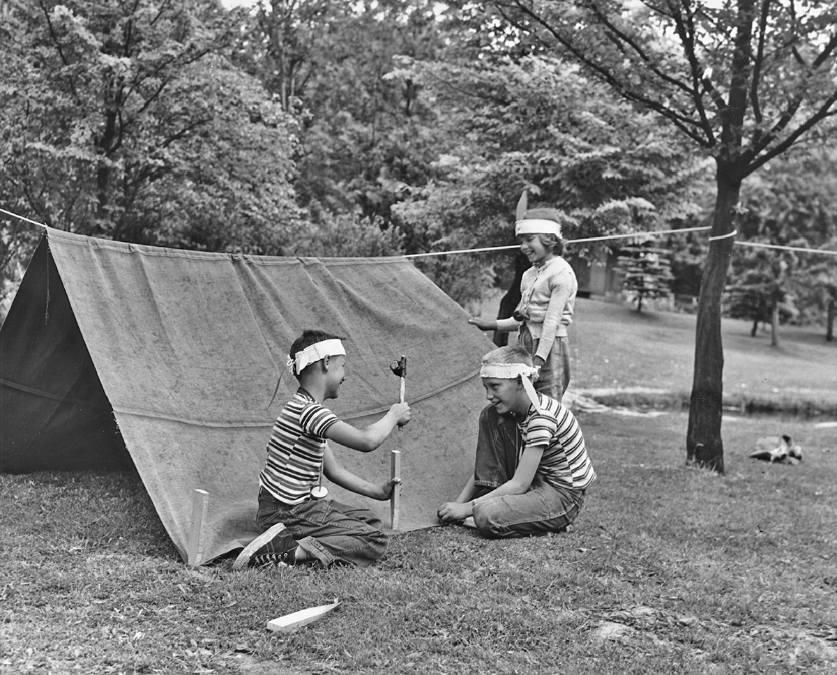 Three children building their own camping tent out of a sheet and rope.