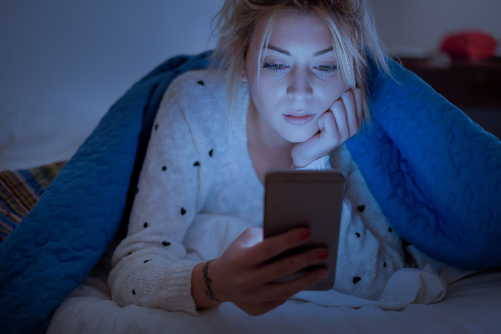 Woman on her phone in bed being kept up because of the blue light.